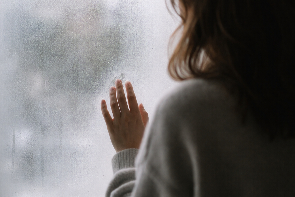Silueta femenina desenfocada de espaldas, de pie frente a una ventana empañada. A través del vidrio se distingue una ciudad borrosa, creando una atmósfera de introspección, pausa y confusión emocional.